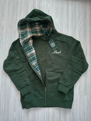 Mörkgrön hoodie med rutigt foder - Stilren mörkgrön hoodie med dragkedja och huva. Insidan av huvan har ett snyggt rutigt foder i grönt, vitt och brunt. Framtill finns fickor och diskret broderad text. Perfekt för en avslappnad look.