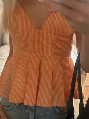 Ny trendig Orange peplumtop med v-ringning - Säljer en orange peplumtop med tunna vita ränder och v-ringad hals. Toppen är ärmlös och har en figurnära passform upptill med utsvängd nederdel. Perfekt för sommarens alla tillfällen!
