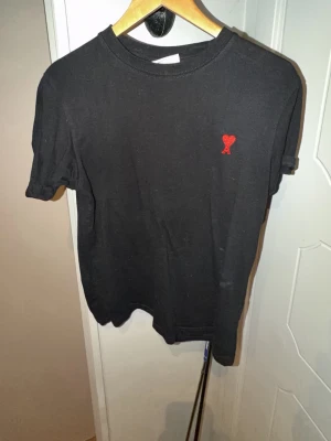 Ami Paris t shirt - Svart t-shirt i bomull med korta ärmar och rund hals. På bröstet finns en liten röd broderad detalj i form av ett hjärta med bokstaven A. Enkel och stilren design som passar till det mesta.
