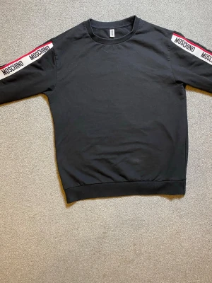 Svart Moschino sweatshirt med logga - Snygg svart sweatshirt från Moschino med vita och röda loggband längs båda ärmarna. Tröjan har rund halsringning, långa ärmar och ribbade muddar vid ärmslut och nederkant. Tillverkad i mjukt bomullsmaterial för en bekväm känsla.