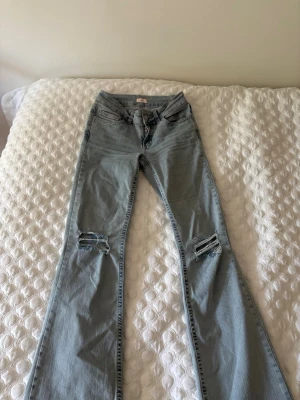 Ljusblå bootcut jeans med slitningar - Säljer ett par ljusblå bootcut jeans med slitna detaljer över knäna och en snygg broderad detalj i rosa på bakfickan. Jeansen har klassisk femficksdesign och är tillverkade i mjukt denimtyg. Perfekta för en avslappnad och trendig look. Byxorna är sparsamt använda
