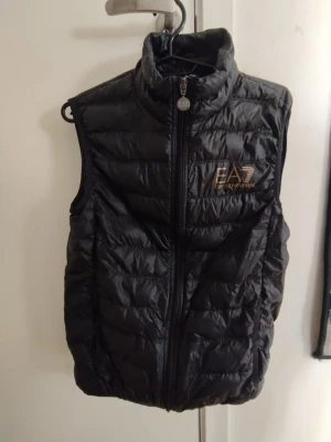 EA7 väst - Snygg svart dunväst från Emporio Armani EA7 med guldfärgad logga på bröstet. Västen har hög krage, dragkedja framtill och quiltad design. Perfekt för lager på lager och ger en sportig look.