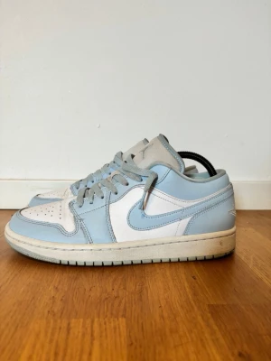 Nike Air Jordan 1 Low blå/vit - Snygga Nike Air Jordan 1 Low sneakers i ljusblått och vitt läder. Klassisk siluett med perforerad tåbox, vita sidopaneler och ljusblå detaljer på häl, snörning och Swoosh. Yttersulan är vit med svart undersida. Perfekta för dig som gillar streetwear och ikoniska sneakers.