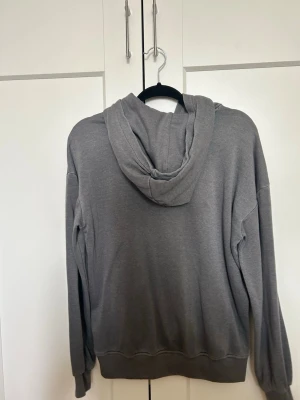 Grå basic hoodie med ficka - En enkel och stilren grå hoodie med huva och stor magficka. Hoodien har långa ärmar, ribbade muddar och avslappnad passform. Perfekt för chill dagar eller när du vill ha något bekvämt och neutralt. Materialet känns mjukt och skönt mot huden.