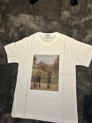 Vit t-shirt med tidningsomslagstryck - Säljer en stilren och snygg T-shirt med tryck från Fiore Studios   T-shirten är vit med ett snyggt vintageinspirerat magasintryck framtill. Enkel att matcha och passar perfekt till både jeans och shorts.  Bilderna / trycket är gjort med presstekniken sublimering. Vilket betyder att trycket färgas i själva fibrerna / trådarna på tyget och INTE som ett plastigt lager ovanpå tyget.   Storlek: S - aldrig använd 