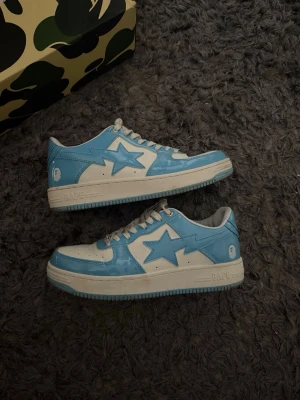 BAPE skor - Säljer ett par BAPE STA sneakers i blått och vitt. Skorna har den ikoniska stjärndesignen på sidan, vita snören och perforerad tåbox. Tillverkade i blankt läder med låg profil och klassisk gummisula. Perfekta för dig som gillar streetwear och vill sticka ut. Storlek 46