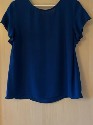 Blå kortärmad blus från VILA - Snygg blå blus från VILA med rund halsringning och korta ärmar med lätt volang. Tillverkad i ett mjukt, tunt tyg som ger ett elegant fall. Perfekt att styla med jeans eller kjol för en trendig look.