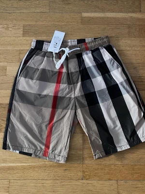 Burberry shorts/ badshorts  - Snygga shorts med klassiskt rutigt mönster i beige, svart, vitt och röda detaljer. Shortsen har elastisk midja med vit snörning och sidofickor. Perfekta för sommaren och har en loose passform. Tillverkade i ett lätt och syntetiskt material.