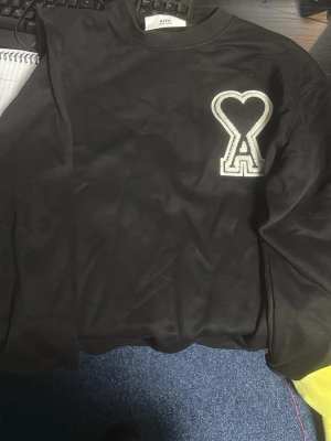 Svart sweatshirt med hjärtdetalj - Säljer en svart sweatshirt från Ami med en stor vit och svart hjärtdetalj och bokstaven A på bröstet. Tröjan har rund halsringning och är tillverkad i mjukt material som känns skönt mot huden. Perfekt för en avslappnad och trendig look.