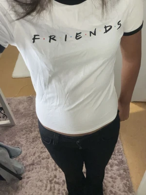 Vit FRIENDS t-shirt med svart kant - Säljer en vit t-shirt med svart kant vid hals och ärmslut. På bröstet står det 'FRIENDS' i svart text med färgglada prickar mellan bokstäverna, inspirerat av den ikoniska tv-serien. T-shirten har en slim passform och är kortärmad. Perfekt för dig som gillar stilrena och klassiska motiv.