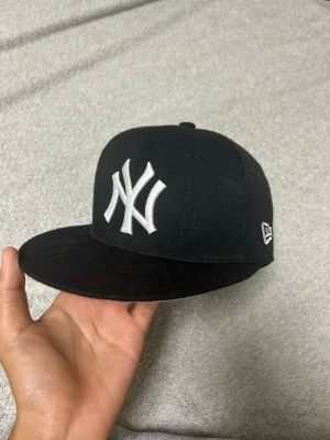 Svart New York Yankees keps - Snygg svart keps från New Era med klassiskt New York Yankees-logga broderad i vitt framtill. Platt skärm och broderad logga på sidan. Perfekt för dig som vill ha en stilren och sportig look.