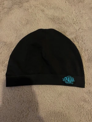 Syna world skull cap - Säljer en svart mössa i mjukt tyg med en liten turkos brodyrdetalj framtill. Enkel och stilren design som passar till det mesta. Perfekt för dig som vill ha en clean look med en liten färgklick.
