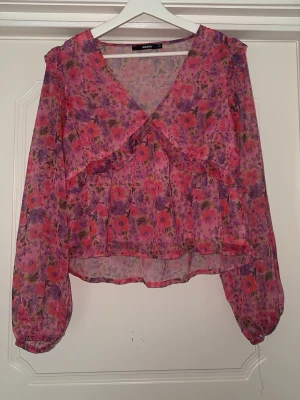 Blommig transparent blus med volanger - Säljer en blommig blus i transparent material med rosa, lila och röda toner. Blusen har volangdetaljer, v-ringning och långa puffiga ärmar med resår vid handlederna. Perfekt för dig som gillar romantisk och färgglad stil.