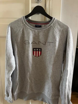 Grå sweatshirt från GANT - Klassisk grå sweatshirt från GANT med rund halsringning och broderad logga samt sköld med stjärnor och ränder på bröstet. Tröjan har långa ärmar och ribbade muddar vid ärmslut och nederkant. Perfekt till en avslappnad stil.