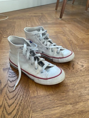 Vita Converse sneakers - Vita Converse sneakers med röd rand runt sulan och vita skosnören. Skorna har gummisula och den ikoniska Converse-loggan på sidan. Säljer nu då de inte passar mig längre❤️