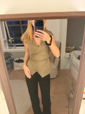 Beige omlottopp med v-ringning - Snygg beige omlottopp med v-ringning och korta ärmar. Toppen har en unik asymmetrisk nederkant och markerad midja med dekorativt spänne i sidan. Perfekt att styla med jeans eller kjol för en trendig look. Prislappen kvar, aldrig använd!