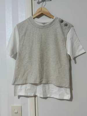 Beige stickad topp med knappar - Trendig beige stickad topp med korta ärmar och dekorativa knappar på axeln. Underdelen är en vit t-shirt som ger en lager-på-lager-look. Perfekt för dig som gillar en avslappnad men ändå stilsäker outfit.
