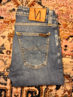 Blå jeans från Nudie Jeans - Säljer ett par klassiska blå jeans från Nudie Jeans med snyggt slitna detaljer och kontrastsömmar. Jeansen har raka ben, två bakfickor med broderad våg och en patch i läder bak i midjan. Perfekta till vardagslooken! Dem sitter slim på och modellen är skinny lin w28 l30