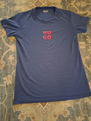 Blå HUGO BOSS t-shirt med tryck - Snygg blå t-shirt från HUGO BOSS med rött HUGO-tryck på bröstet. Klassisk rund hals och korta ärmar. Tillverkad i mjukt material som känns skönt mot huden. Perfekt för en avslappnad och stilren look.