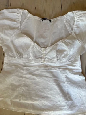 Brandy topp!! - Söt vit blus med puffiga korta ärmar och djup urringning. Blusen har dekorativa knappar framtill och markerad midja. Tillverkad i ett lätt, vävt material som ger en romantisk känsla. Den är one size! 