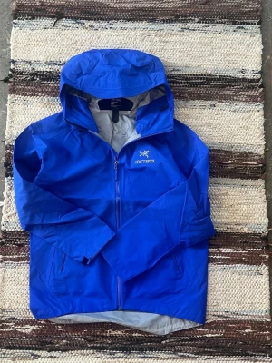 Blå regnjacka från Arc'teryx - Snygg blå regnjacka från Arc'teryx med huva och hel dragkedja framtill. Jackan har två sidofickor med dragkedja och är tillverkad i ett lätt, vattentätt material. Perfekt för regniga dagar och outdoor-äventyr.