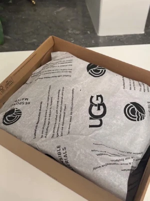 Mörkbruna UGG tofflor i mocka - Säljer ett par klassiska UGG tofflor i mörkbrun mocka med mjukt och fluffigt foder. Tofflorna har en rundad tå och låg ankelhöjd, perfekta för att hålla fötterna varma och sköna. Ikonisk UGG-design och riktigt mysiga att ha på sig.
