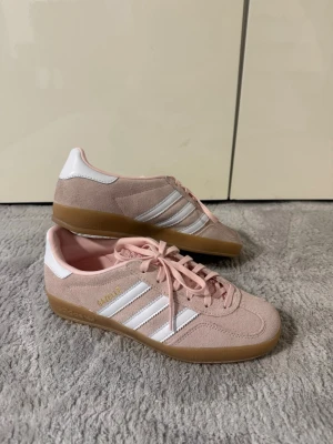 Adidas Gazelle rosa sneakers - Snygga Adidas Gazelle sneakers i ljusrosa mocka med vita detaljer och klassiska tre ränder på sidorna. Skorna har vit hälkappa, rosa skosnören och en gummisula i brunt.