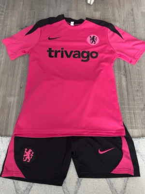 Chelsea träningsset rosa/svart Nike S - Chelsea träningsset från Nike i rosa och svart. Tröjan har korta ärmar, svart axelparti och tryck med klubbmärke och sponsor. Matchande shorts med rosa detaljer och klubbmärke. Tillverkad i lätt, ventilerande material perfekt för träning.