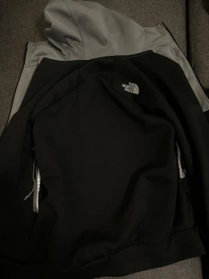 Svart vindjacka från The North Face - Snygg svart vindjacka från The North Face med grå huva och vita detaljer vid dragkedjorna. Jackan har en klassisk logga på bröstet och praktiska fickor med dragkedja. Perfekt för dig som vill ha en stilren och funktionell jacka.