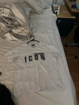 Vit ICON t-shirt från Dsquared2 - Storlek L men sitter som M Vit t-shirt från Dsquared2 med trycket 'ICON' i svart på bröstet. Klassisk rund hals och korta ärmar. Perfekt för dig som gillar streetwear och vill sticka ut med en ikonisk logga. Tröjan är lite använd och har ett litet hål längst upp på baksidan inget man märker dock