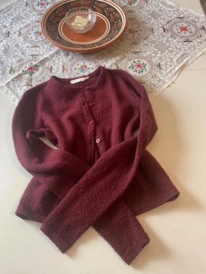 Vinröd kofta från Uniqlo U - Mysig vinröd kofta från Uniqlo U med rund halsringning och knäppning framtill. Tillverkad i ett mjukt, strukturerat material som ger en avslappnad look. Perfekt att slänga över en t-shirt eller klänning för extra värme.Vinröd höst kofta 2000s i storlek ungefär xxs perfekt för hösten och vintern ifrån Uniq studio.
