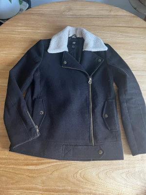 Svart teddyjacka från H&M - Säljer en svart teddyjacka från H&M med vit, fluffig krage i fuskpäls. Jackan har sned dragkedja, dekorativa knappar och fickor med lock. Materialet är mjukt och värmande, perfekt för kyliga dagar. Snygg bikerstil som ger en cool look.