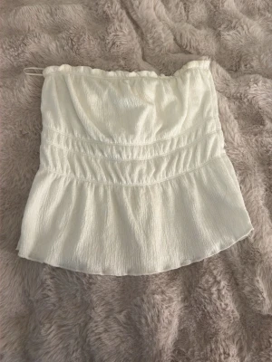 Vit axelbandslös tubtopp - Säljer en söt vit tubtopp med lätt rynkad struktur och volang nedtill. Toppen är axelbandslös och har en mjuk, flowy passform som passar perfekt till sommaren. Perfekt att matcha med jeans eller kjol för en fräsch look.