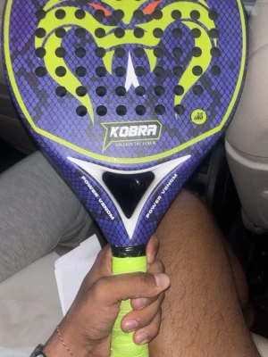 Kobra padelracket med ormmönster - Säljer ett padelracket från Kobra med coolt ormmönster i lila, svart och neongrönt. Racketen har hål i träffytan och grepp i neongrönt. Perfekt för dig som vill sticka ut på padelbanan.