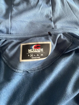 Blå hoodie från McKenzie med dragkedja - Snygg blå hoodie från McKenzie med dragkedja framtill och huva. Märkeslogga på bröstet och vita detaljer längs axlarna. Tillverkad i ett mjukt och stretchigt material som känns skönt mot huden. Perfekt för en avslappnad och sportig stil. Köpt på JD sports i butik!