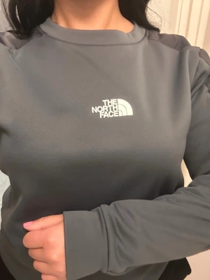 Grå sweatshirt från The North Face - Snygg grå sweatshirt från The North Face med rund halsringning och diskret logga på bröstet. Tröjan har mörkare partier på axlar och armbågar samt långa ärmar. Perfekt för en avslappnad och sportig stil.