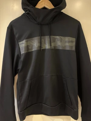 Svart hoodie från Jordan - Säljer en svart hoodie från Jordan med camo-inspirerad detalj och text över bröstet. Hoodien har stor huva med dragsko och en rymlig magficka. Tillverkad i mjukt material som känns skönt mot huden. Perfekt för dig som gillar streetwear och sportig stil.