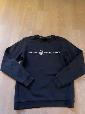 Blå Sail Racing sweatshirt - Cool blå sweatshirt från Sail Racing med vit logga tryckt på bröstet. Tröjan har rund hals, långa ärmar och ribbade muddar vid ärmslut och nederkant. Perfekt för en avslappnad och sportig stil.