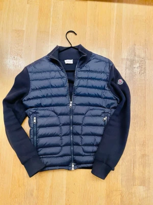 Mörkblå cardiganjacka från Moncler - Snygg mörkblå cardiganjacka från Moncler med quiltad framsida och stickade ärmar. Jackan har dragkedja framtill, två fickor med dragkedja och Moncler-logga på ärmen. Perfekt för lager-på-lager och ger en sportig men stilren look.