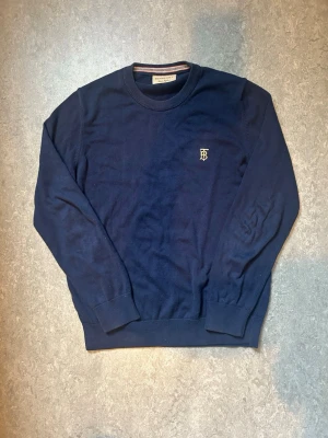 Mörkblå tröja med broderad logga - Snygg mörkblå tröja med rund hals och ribbade muddar. På bröstet finns en liten broderad logga i gult som ger en exklusiv känsla. Perfekt till jeans eller chinos för en clean look.