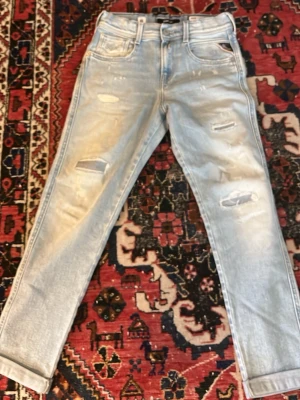Ljusblå slitna  jeans - Säljer ett par ljusblå mom jeans med slitningar och hål på framsidan. Jeansen har fem fickor och uppvikta benslut. Perfekta för en avslappnad och trendig look. Materialet är klassisk denim i bomull.