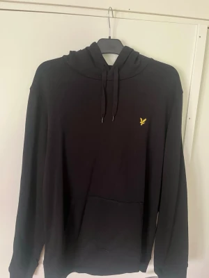 Svart hoodie med gul logga - Snygg svart hoodie med huva och dragsnören samt en gul broderad logga på bröstet. Klassisk känguruficka framtill och ribbade muddar vid ärmar och nederkant. Perfekt för en chill och avslappnad stil.