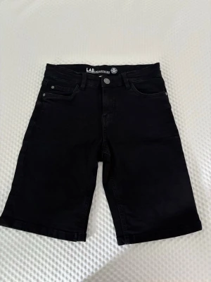 Svarta jeansshorts från LAB - Snygga svarta jeansshorts från LAB med klassisk femficksmodell och knappgylf. Shortsen har raka ben och är tillverkade i ett slitstarkt denimtyg. Perfekta för en avslappnad och trendig stil.