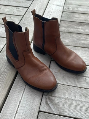 Bruna Chelsea boots i läder - Snygga bruna Chelsea boots i slätt läder med svarta elastiska paneler på sidorna och dragkedja på insidan. Klassisk rund tå och låg klack. Perfekta för dig som vill ha en stilren och tidlös look till hösten.