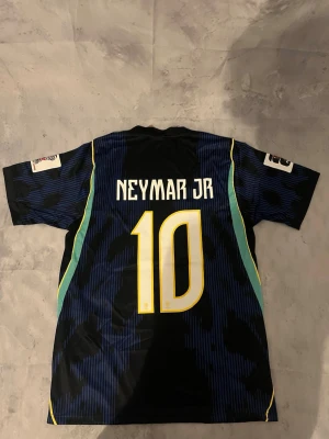 Brasilien Neymar JR matchtröja blå - Snygg blå matchtröja från Brasilien med Neymar JR och nummer 10 på ryggen. Tröjan har gula och gröna detaljer, korta ärmar och officiella tryck på bröstet och ärmarna. Perfekt för dig som älskar fotboll och vill stötta landslaget.