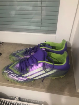Lila och limegröna F50 fotbollsskor - Säljer ett par snygga F50 fotbollsskor i lila och limegrönt med vita detaljer. Skorna har snörning, låg profil och dobbar för bra grepp på planen. Insidan är limegrön och ovandelen har en glansig finish med coola mönster. Perfekta för dig som vill sticka ut på fotbollsplanen.