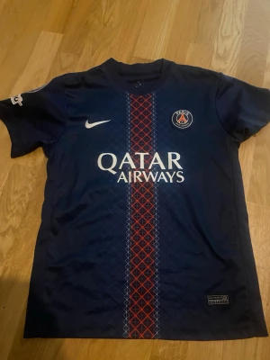 PSG matchtröja blå Nike - Säljer en blå PSG matchtröja från Nike med Qatar Airways-tryck på bröstet och klubbmärke. Tröjan har ett rött och blått mönster längs mitten och nummer 14 med namnet D. Doué på ryggen. Perfekt för fotbollsfans!