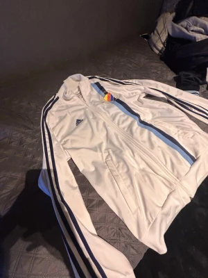 Vit Adidas träningsjacka med blå ränder - Säljer en vit träningsjacka från Adidas med blå ränder längs ärmarna och en DIF-logga på bröstet. Jackan har dragkedja framtill, ståkrage och två fickor. Perfekt för sport eller chill. Materialet är troligtvis polyester.