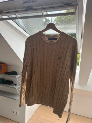 Beige kabelstickad tröja Polo Ralph Lauren - Säljer en beige kabelstickad tröja från Polo Ralph Lauren med rund halsringning och den klassiska broderade loggan på bröstet. Tröjan har ribbade muddar vid ärmslut och nederkant. Perfekt för dig som gillar stilrena och tidlösa plagg. Storlek S men passar även M. 