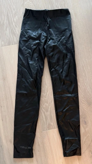 Svarta glansiga leggings - Säljer ett par svarta leggings med glansig finish och hög midja. De har en tight passform och är tillverkade i ett stretchigt material som ger en cool look. Perfekta att styla med oversized tröja eller crop top.